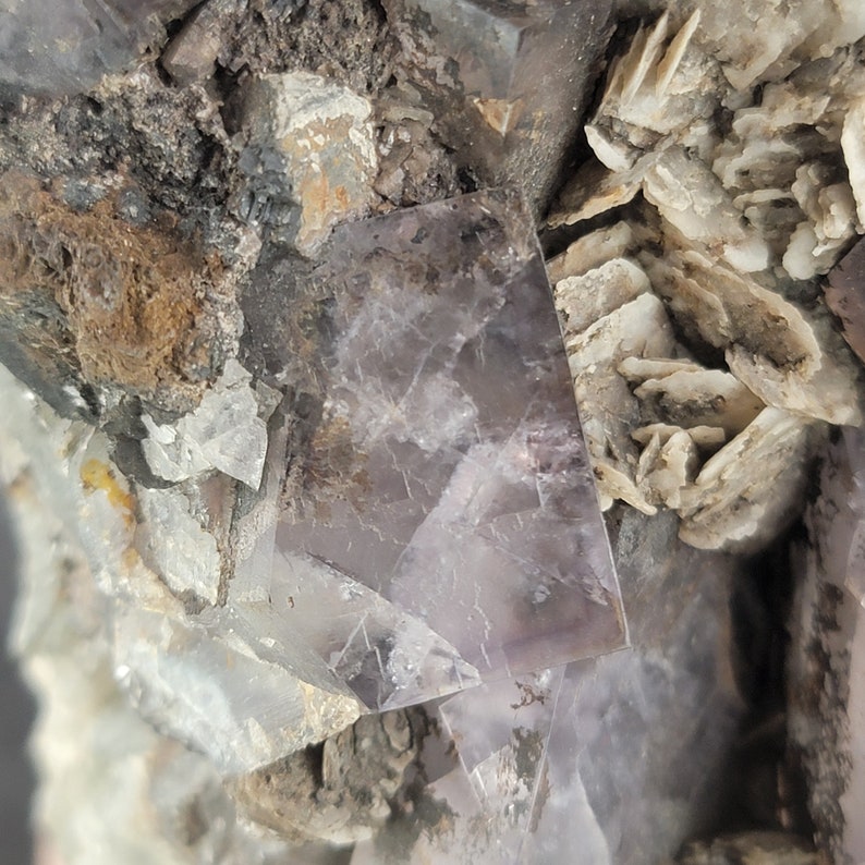 Op de afbeelding: Een close-up van een paarse fluorietkristal met een ruw, getextureerd oppervlak. De kristal is omgeven door andere kleinere kristallen en een wit, gelaagd mineraal.