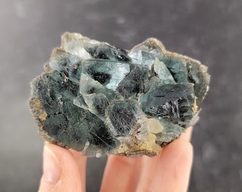 Pode incluir: Um aglomerado de cristais de fluorita verde com uma variação de cor verde escuro e verde claro. Os cristais estão agrupados em uma formação natural áspera.