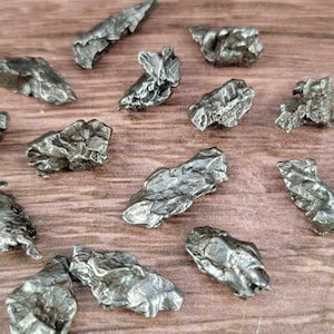 Könnte beinhalten: Eine Sammlung von 13 kleinen, unregelmäßig geformten, silberfarbenen Meteoritenfragmenten auf einer braunen Holzoberfläche.