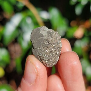 May include: A close-up of a small, rough, gray and silver rock with a sparkly, metallic texture. The rock is being held in a person's hand.