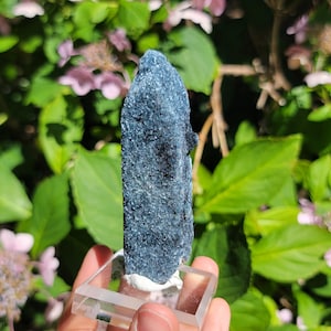 May include: A dark blue, textured mineral specimen with a crystalline, sparkling surface. The elongated, vertical stone is mounted upright on a clear, square acrylic base, showcasing a rough, natural formation with deep blue and black tones.