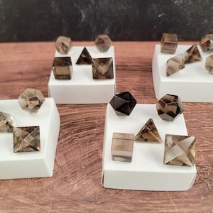 May include: Smoky quartz crystals in various geometric shapes, including cubes, pyramids, and multi-faceted forms, presented on white platforms. The crystals are a translucent brown colour.