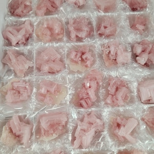 Puede incluir: Una colección de grupos de cristales rosas, cada uno envuelto individualmente en plástico transparente. Los cristales tienen una apariencia suave y translúcida y están dispuestos en un patrón de cuadrícula, lo que sugiere una exhibición de especímenes minerales.