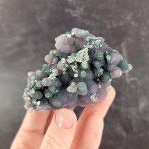 May include: A cluster of grape agate, a unique gemstone with a bubbly, botryoidal structure. The stone features a mix of purple, lavender, and green hues, with a textured surface. The gemstone is held in a hand.
