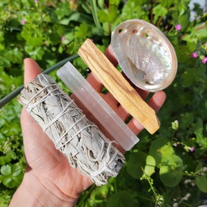 May include: A collection of spiritual items including a white sage bundle, a clear selenite wand, a palo santo stick, and an iridescent abalone shell. The items are arranged in a hand against a backdrop of green foliage.