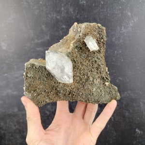 May include: A cluster of clear quartz crystals embedded in a rough, gray, and brown rock matrix. The crystals are sharp and have a glassy appearance.