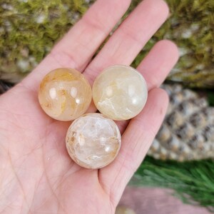 Puede incluir: Tres esferas de cristal de cuarzo pulidas, lisas y transparentes con un tono amarillo dorado. Las esferas se muestran en una mano.