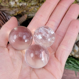 May include: Three clear quartz crystal spheres of varying sizes, displayed on a hand.