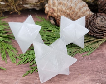 Puede incluir: Tres puntas de estrella de selenita blanca, también conocidas como estrella de seis puntas o hexagrama, están dispuestas sobre una superficie de madera con follaje verde.