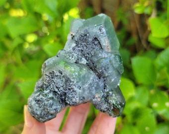 Könnte beinhalten: Ein Cluster aus grünen und grauen Kristallen mit einer rauen, natürlichen Textur. Die Kristalle sind in einer einzigartigen Formation zusammengefügt.
