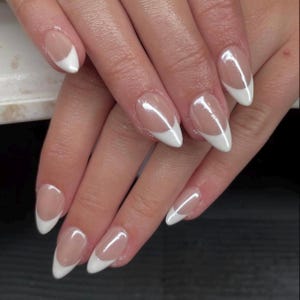 Può includere: Primo piano di una mano con unghie a mandorla. Ogni unghia presenta una base trasparente con una punta bianca, creando un classico look da manicure francese. Le unghie sono lucide e ben curate.