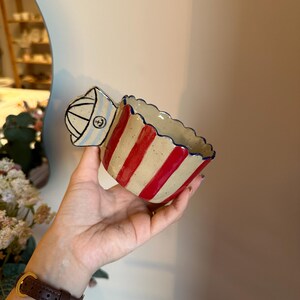 May include: A ceramic bowl with a nautical theme. The bowl has alternating red and beige stripes, a scalloped rim, and a handle shaped like a sailor's hat with an anchor emblem. The bowl is held by a person.
