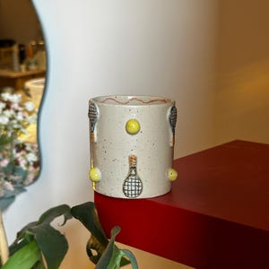 May include: A cylindrical ceramic mug with a speckled off-white glaze, decorated with raised, hand-painted tennis rackets and bright yellow tennis balls. The rim features a thin, wavy red line detail, and the surface has a textured, artisanal finish.