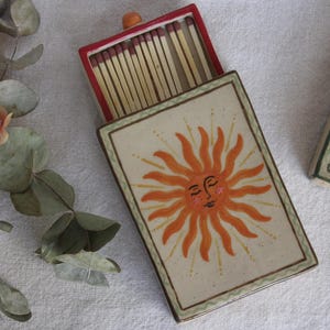 May include: A ceramic matchbox with a hand-painted orange sun design. The matchbox is rectangular with a red interior and a lid. The sun has a smiling face. The image also shows a sprig of eucalyptus and flowers.