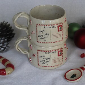 May include: Two stacked ceramic mugs with twisted handles, decorated with candy canes and postcard designs that read "Have a Holly Jolly Christmas!". The scene includes Christmas ornaments, pine cones, and a candy cane spoon.