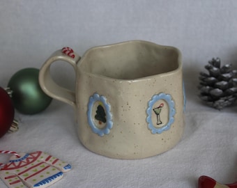 May include: A speckled beige ceramic mug with a square shape and a curved handle. The mug features blue framed illustrations of a Christmas tree and a cocktail. Christmas ornaments and pine cones are in the background.