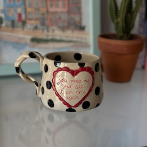 Könnte beinhalten: Cremefarbene Keramiktasse mit schwarzen Tupfen und einem gedrehten Henkel. Ein rotes Herz mit der Aufschrift "You make me feel like I am home again" ist mittig auf der Vorderseite. Ein Kaktus und ein Gemälde sind im Hintergrund.