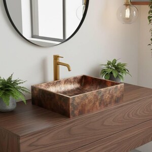 May include: A modern bathroom vanity with a rectangular copper sink and a gold faucet. The dark wood cabinet has two drawers and is mounted on the wall. A round black mirror hangs above the sink, and plants add a touch of greenery.