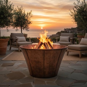Può includere: Un braciere in rame con fuoco acceso, posto su un patio in pietra. Il braciere è circondato da mobili da esterno e alberi, con un tramonto sul mare sullo sfondo.