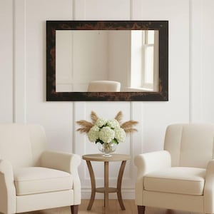 May include: A rectangular mirror with a dark brown, textured frame hangs above two cream-colored armchairs. A small round table with a floral arrangement sits between the chairs. The wall is white with vertical paneling.