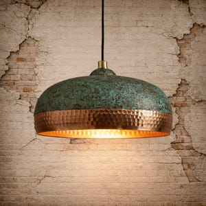 May include: A hanging pendant light with a copper and verdigris finish. The dome-shaped shade has a textured, aged appearance with a copper band. The light fixture hangs from a black cord against a brick wall.