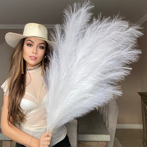 Può includere: Una donna con un cappello bianco e una camicetta bianca tiene in mano un grande pennacchio di erba della pampa bianca. Il pennacchio è molto soffice e ha un aspetto naturale e secco.