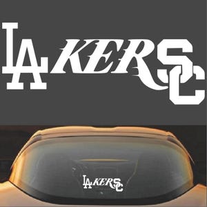 May include: White vinyl decal with the text "LAKERS" in a stylized font. The "LA" is in a traditional font and the "SC" is in a stylized font. The decal is shown on the back window of a car.