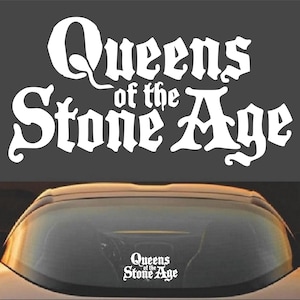 May include: White vinyl sticker with the text "Queens of the Stone Age" in a gothic font. The sticker is on the rear window of a car.