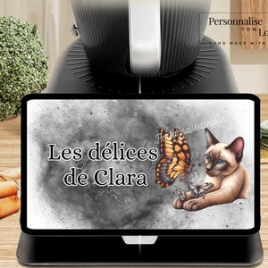 May include: A black tablet displaying the text "Les délices de Clara" with a butterfly and a Siamese cat illustration. The tablet is on a black stand, with a black appliance in the background. Broccoli and carrots are also visible.