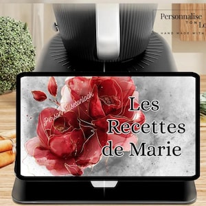 May include: A black kitchen appliance with a white handle is in the background. In the foreground is a black tablet displaying a watercolor image of a red rose with gold accents. The text "Les Recettes de Marie" is displayed on the tablet.