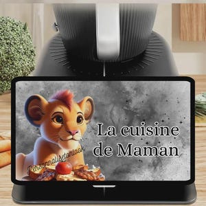 May include: A black digital display featuring a cartoon lion cub with the text "La cuisine de Maman". The display is set on a black base, with a blurred background of a kitchen appliance and vegetables.