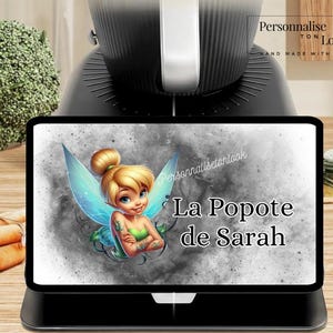 May include: A black digital tablet displays a cartoon fairy with blonde hair, green top, and blue wings. The text "La Popote de Sarah" is written in white. The tablet is on a black stand, with a black appliance in the background.