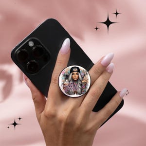 May include: A black mobile phone with a pop-up grip. The grip displays a woman with grey hair, a black cap, and the words "LET THEM". The phone is held in a hand with long, manicured nails.