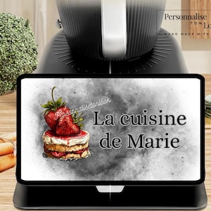 May include: A black kitchen appliance with a digital display featuring a strawberry cake illustration and the text "La cuisine de Marie." The display is set on a wooden surface, with a broccoli floret and carrots visible.