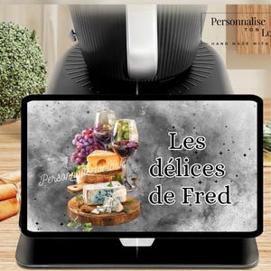 May include: A black tablet displays a food and drink image with the text "Les délices de Fred." The image features wine glasses, grapes, and cheese on a wooden board. The tablet is on a black stand.