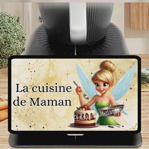 May include: A black kitchen appliance with a digital display showing a cartoon fairy holding a spoon and a bowl. The display reads "La cuisine de Maman" with a cake illustration.
