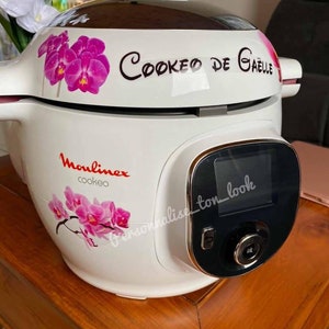 Peut inclure: Cookeo blanc avec un écran digital noir et une bordure argentée. Le cookeo a un motif floral avec des orchidées roses et le texte "Cookéo de Gaëlle" sur le couvercle. La marque "Moulinex" et "cookéo" sont imprimées sur le devant du cookeo.