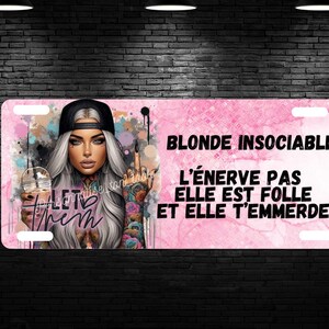 Puede incluir: Placa de matrícula con fondo rosa y texto "BLONDE INSOCIABLE L'ÉNERVE PAS ELLE EST FOLLE ET ELLE T'EMMERDE!". La placa presenta una ilustración de una mujer con tatuajes, gorra negra y bebida.