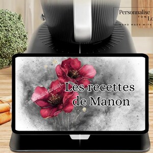 May include: A black tablet displays the text "Les recettes de Manon" over a watercolor floral design. The tablet is on a black stand, with a black kitchen appliance in the background. Broccoli and carrots are also visible.