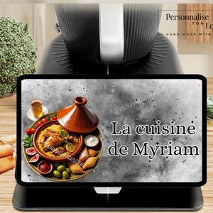 May include: A black kitchen appliance with a digital display showing a plate of food, including a tagine, olives, and tomatoes. The display reads "La cuisine de Myriam."