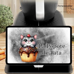 May include: A black digital tablet displays a cartoon cat on a cupcake with the text "La Popote de Tata". The cat is gray and white with a red strawberry on its head. The cupcake has chocolate frosting and sprinkles.