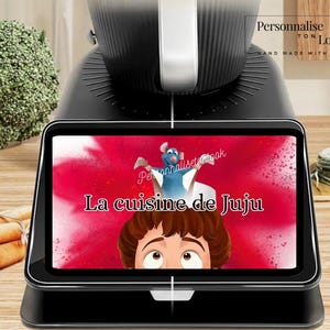 May include: A black kitchen appliance with a digital display showing a cartoon image of a rat and a boy with the text "La cuisine de Juju". The display is set against a red background. The appliance is on a wooden surface.