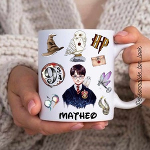 Può includere: Tazza in ceramica bianca con illustrazioni a tema Harry Potter, tra cui un cappello parlante, un gufo e lo stemma di Hogwarts. La tazza ha il nome "MATHEO" stampato sopra. La tazza è tenuta da una persona.