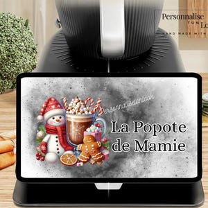 May include: A black tablet displaying a Christmas-themed illustration with a snowman, hot chocolate, and gingerbread man. The text "La Popote de Mamie" is visible. The tablet is on a black stand.