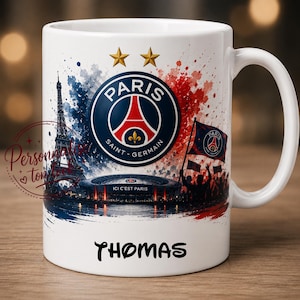 Könnte beinhalten: Weiße Keramik-Tasse mit einem Design, das das Logo von Paris Saint-Germain, den Eiffelturm und ein Stadion zeigt. Die Tasse hat einen Henkel und den Namen "THOMAS" aufgedruckt. Das Design enthält die Farben Rot, Weiß und Blau.