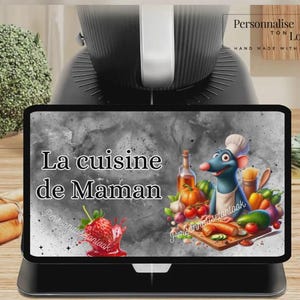 May include: A black tablet displays the text "La cuisine de Maman" with a cartoon illustration of a chef rat and vegetables. The tablet is on a black stand, with a black kitchen appliance in the background.