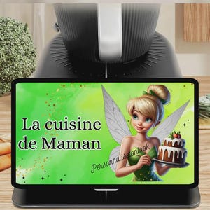 May include: A black digital kitchen appliance with a screen displaying the text "La cuisine de Maman" and a cartoon fairy holding a cake. The screen has a green background with a cartoon fairy holding a cake.
