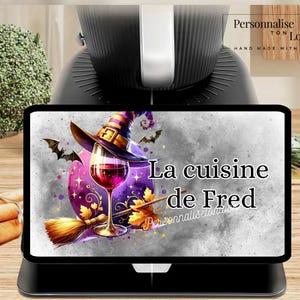 May include: A black tablet displays a colorful graphic with a witch's hat, wine glass, and broom. The text "La cuisine de Fred" is featured. The tablet is on a black stand, with a kitchen appliance in the background.