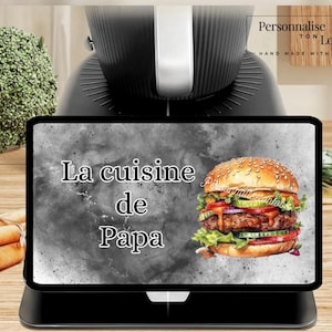 May include: A black digital tablet displays a colorful illustration of a burger with the text "La cuisine de Papa" in white. The tablet is on a black stand, with a black appliance in the background.
