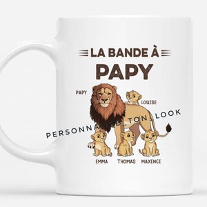 Peut inclure: Mug en céramique blanche avec une illustration de famille de lions. Le texte "LA BANDE À PAPY" est au-dessus de l'illustration du lion, avec des noms sous les lionceaux. La tasse a une anse incurvée.
